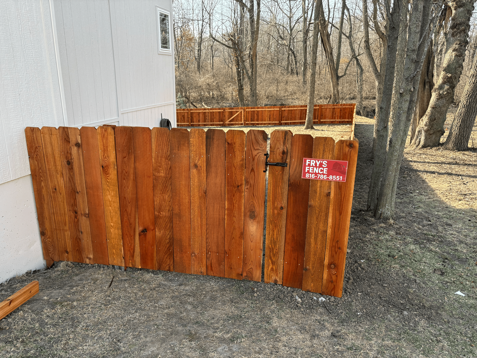 Rustic wooden fence panels stacked beside a white building in a wooded yard.