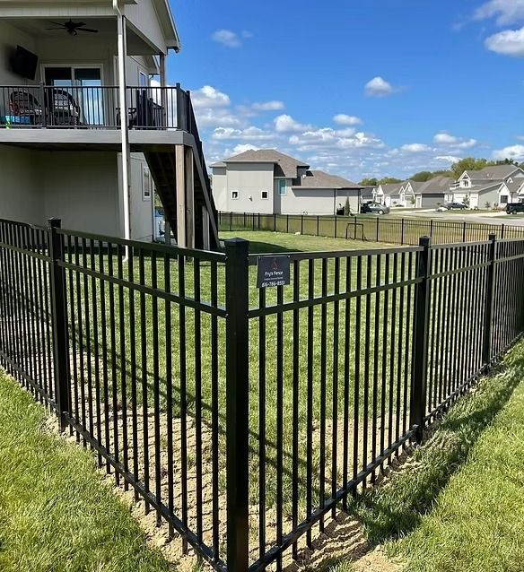 Black metal fence corner in a grassy backyard with suburban houses and a blue sky behind.