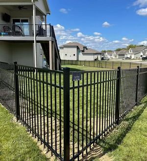 Black metal fence corner in a grassy backyard with suburban houses and a blue sky behind.