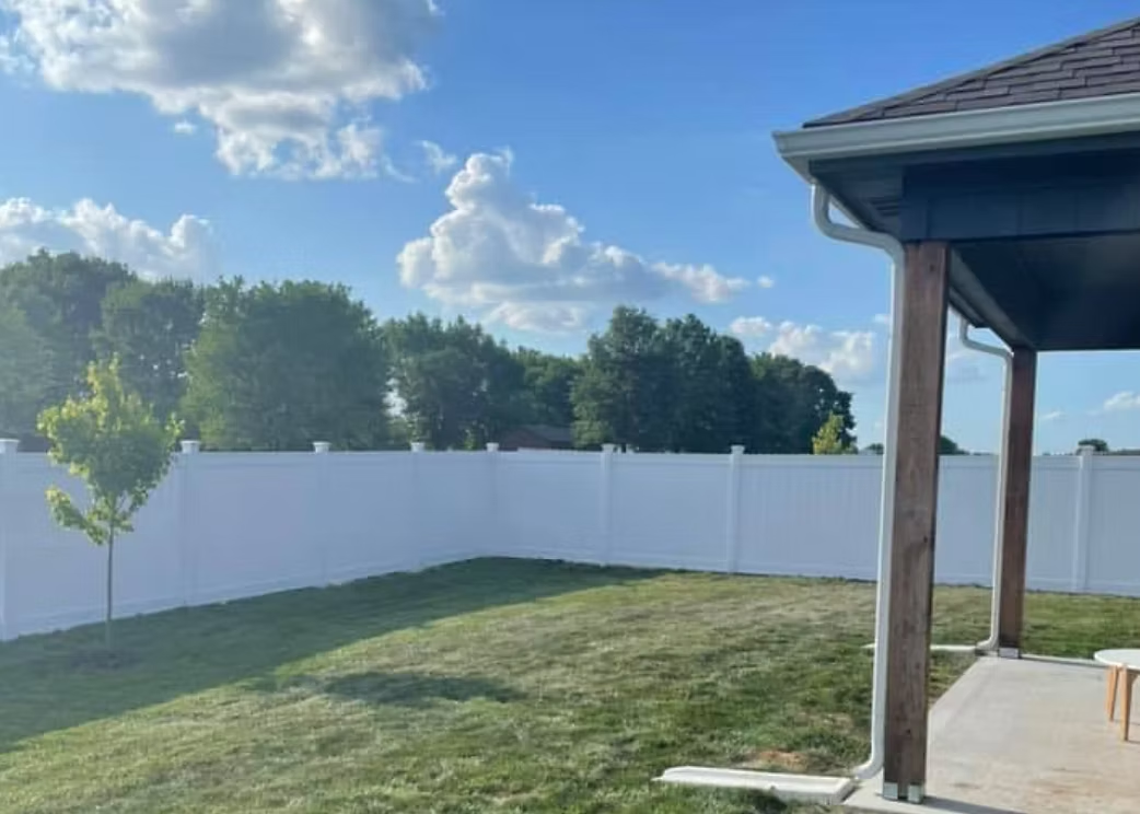 Backyard with white fence, green lawn, trees, and a covered patio under a blue sky