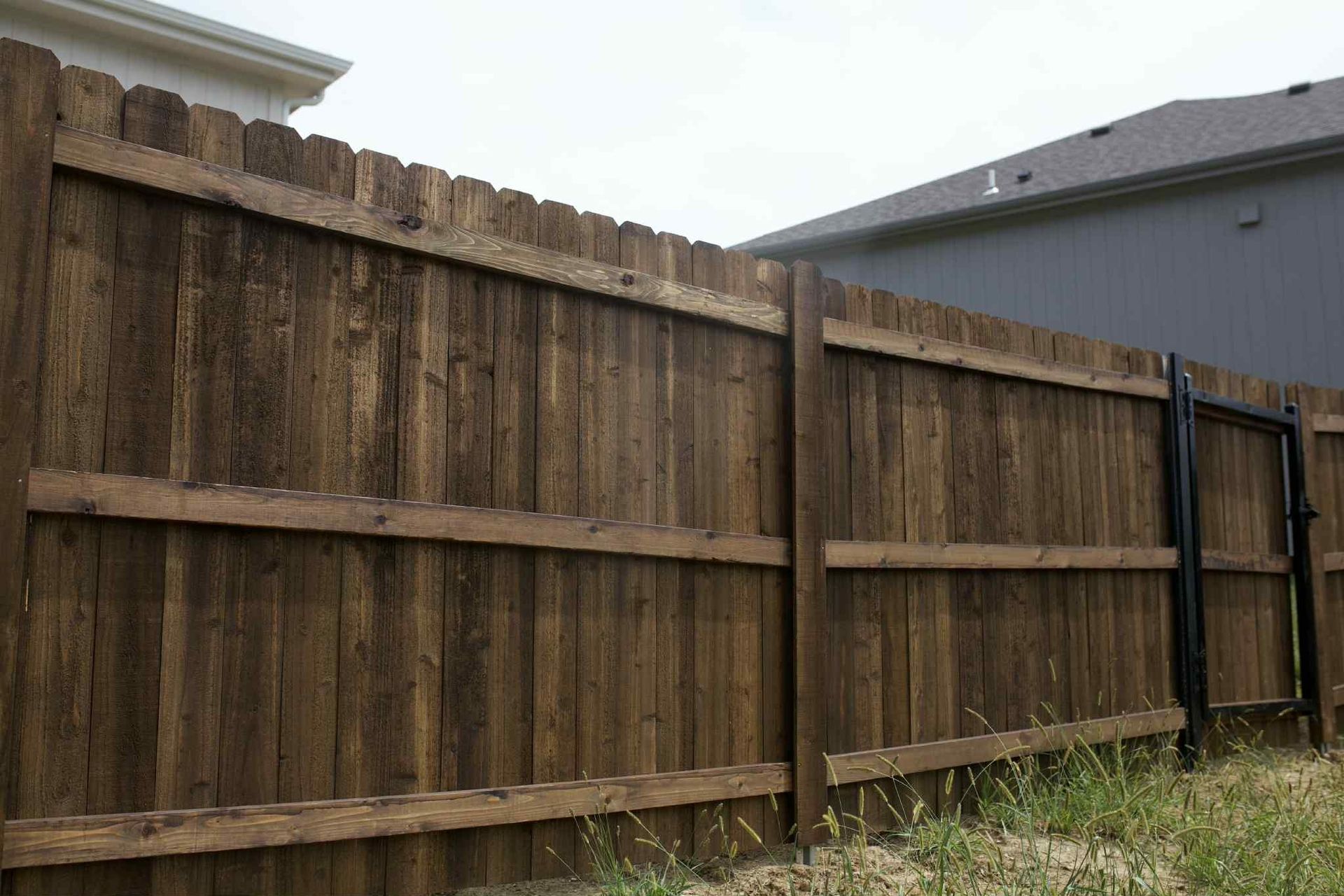 Weathered wooden backyard fence with horizontal supports beside a gray house and grassy yard