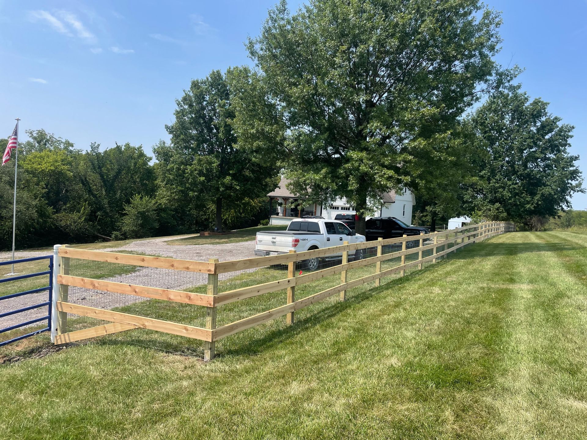 Rural house behind a wooden fence, with a white car and an American flag in a sunny green yard.