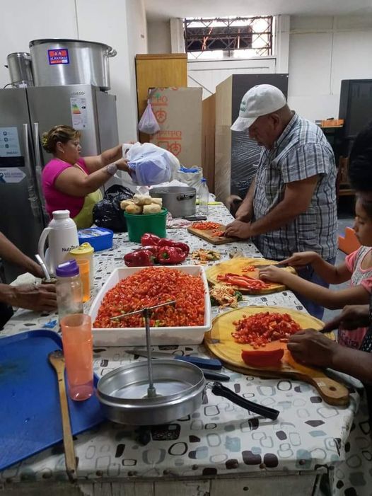 Gente preparando comida en una mesa de cocina abarrotada de bandejas con ingredientes picados y botellas.