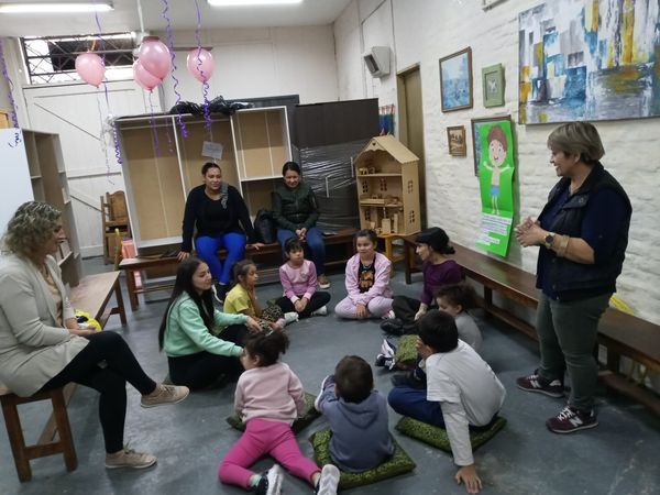 Adultos y niños sentados en un aula, escuchando a una mujer que habla junto a una colorida pared decorada con murales.