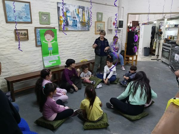 En un aula, los niños están sentados en círculo en el suelo, escuchando a dos adultos que hablan cerca de la pared.