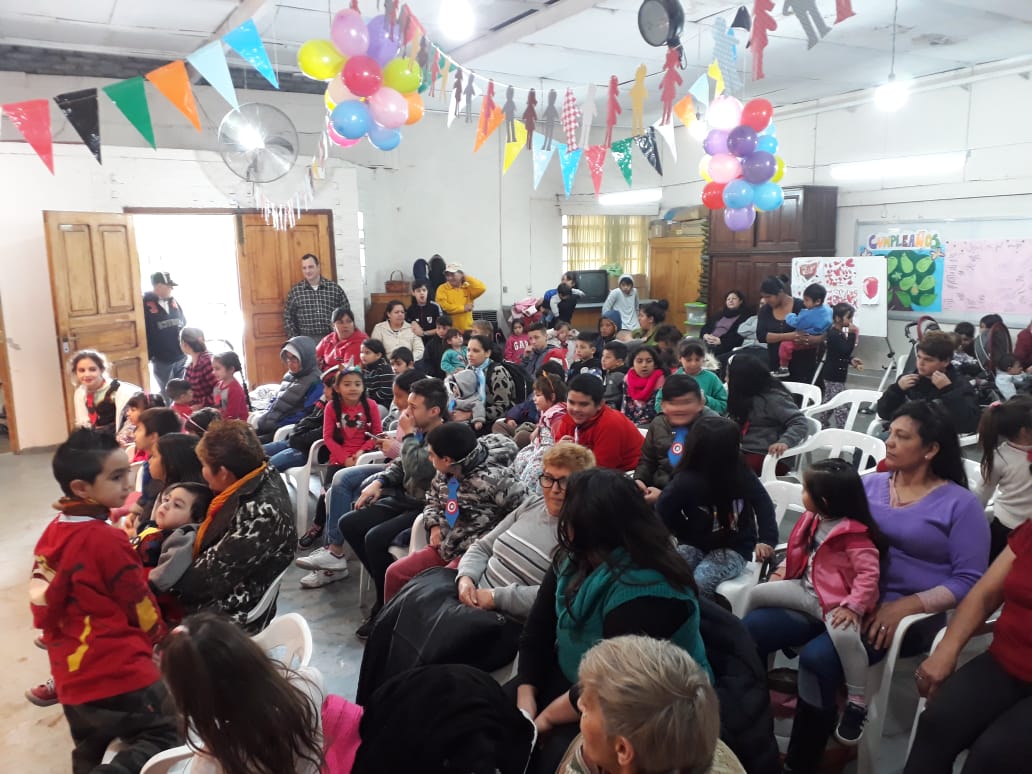 Sala de fiestas abarrotada de gente sentada, globos y guirnaldas de colores en lo alto, y niños con ropa festiva.