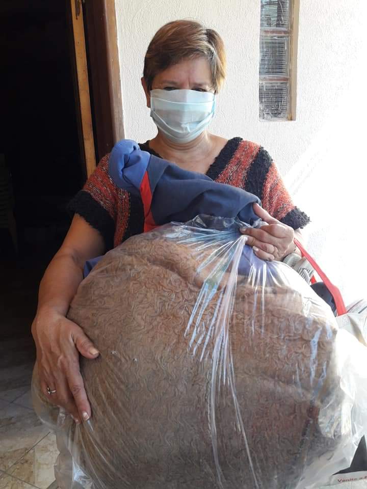 Una persona con mascarilla y delantal sostiene una bolsa grande de grano o harina en el interior de un edificio.