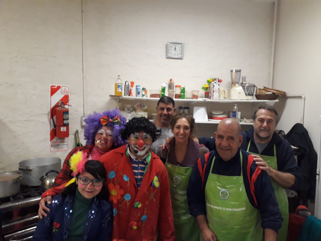 Grupo de personas sonrientes con coloridos disfraces y delantales posando en una cocina con estantes y utensilios de cocina.