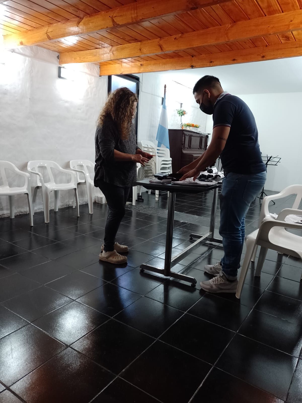 Dos personas están de pie junto a una mesa pequeña en una habitación blanca con suelo de baldosas negras y sillas de plástico.