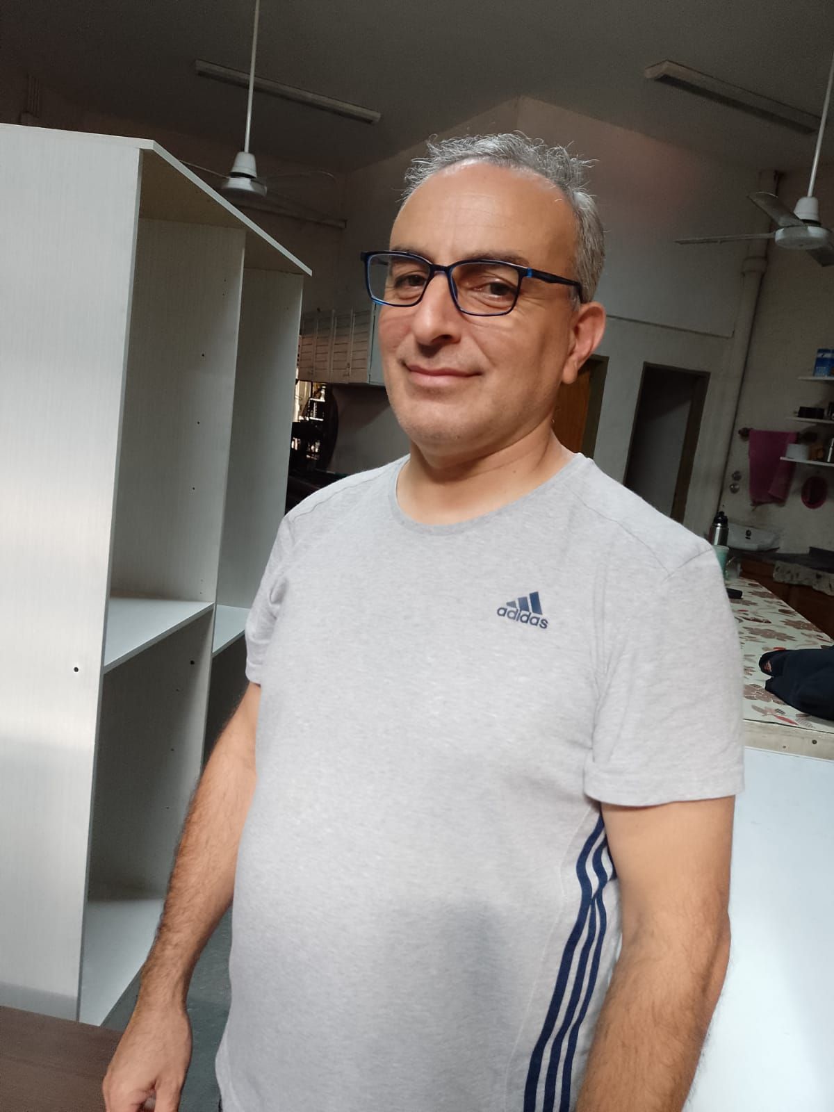 Hombre con gafas y camiseta Adidas gris claro de pie en el interior de una habitación con estanterías y ventiladores de techo.
