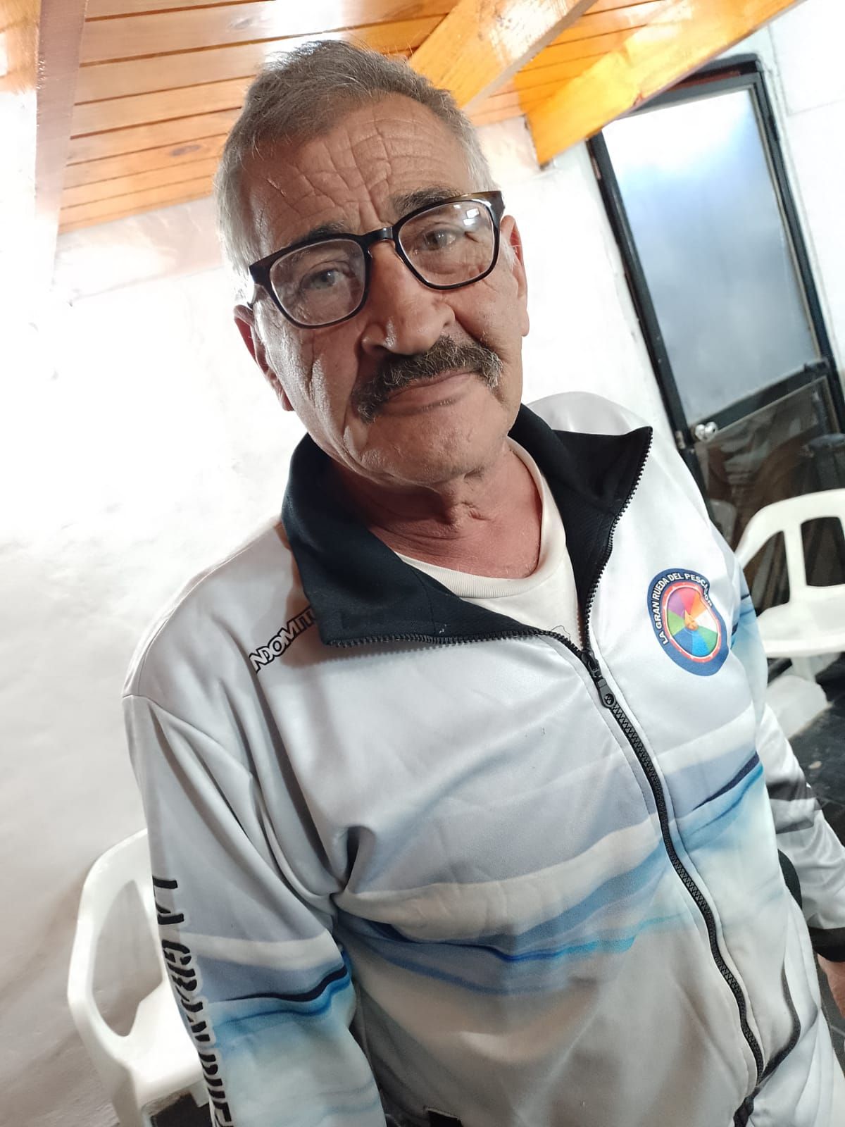Hombre con gafas y chaqueta blanca con cremallera, sentado en el interior de una vivienda, apoyado contra una pared de azulejos.