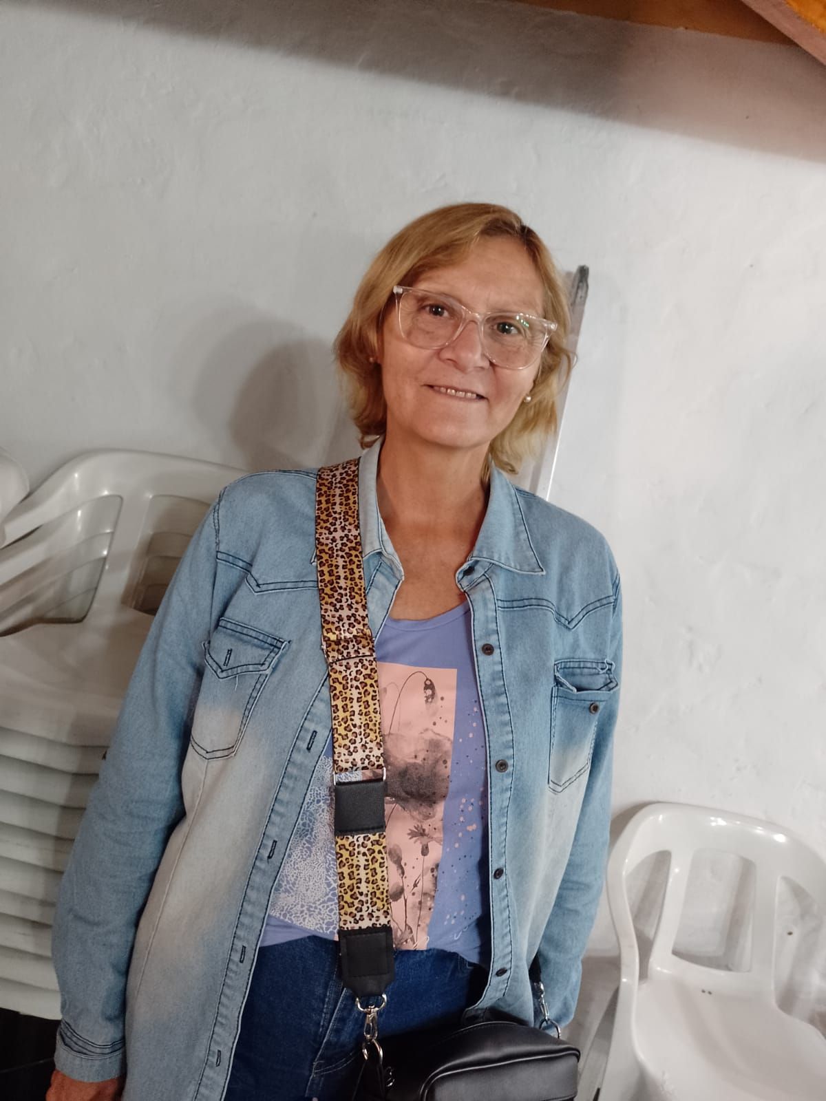 Mujer de pie en el interior de una vivienda, apoyada contra una pared blanca, con gafas, una chaqueta vaquera y una bandolera estampada.