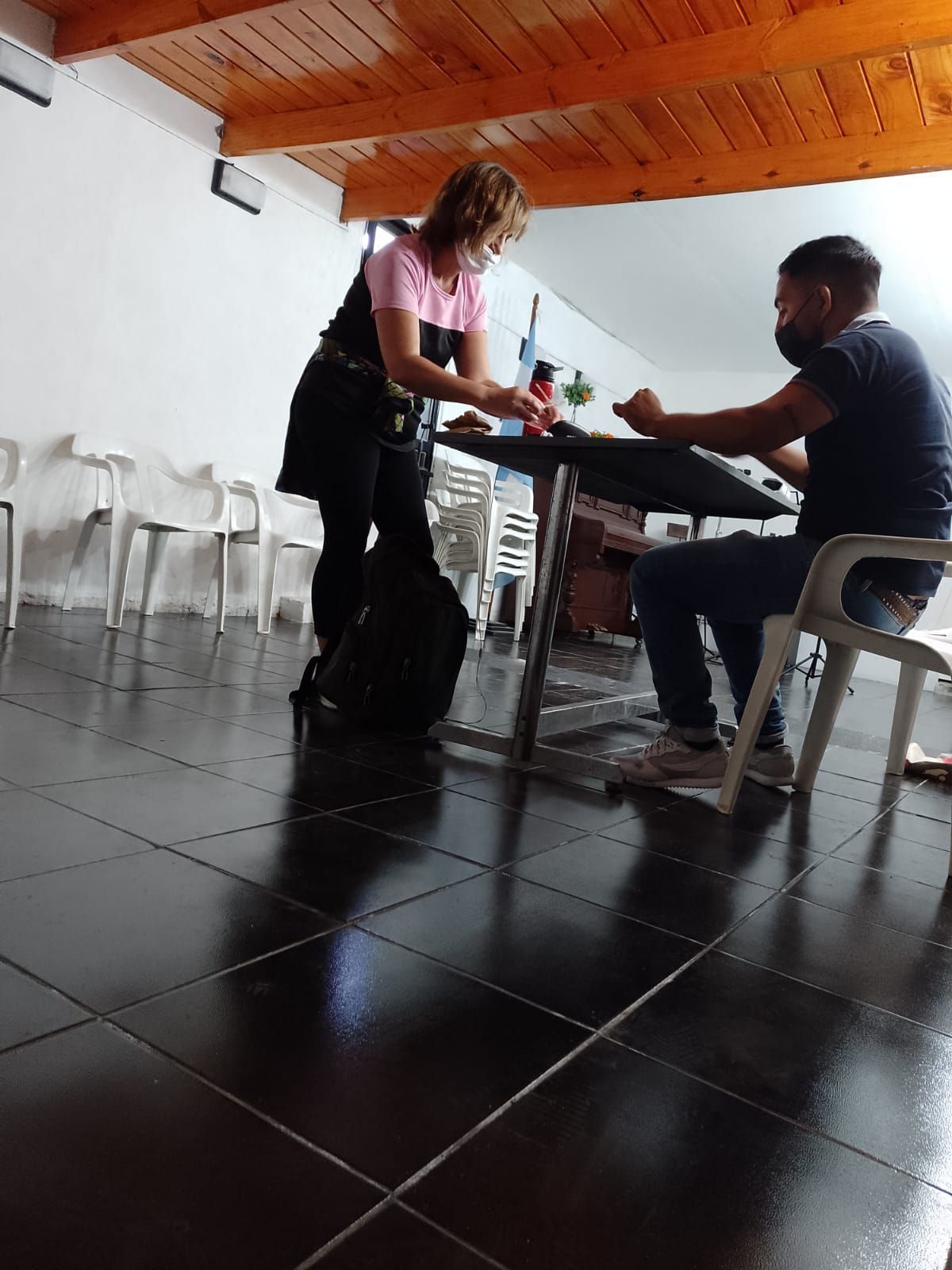 Dos personas sentadas a una mesa en una habitación con suelo de baldosas; una le entrega una flor a la otra.