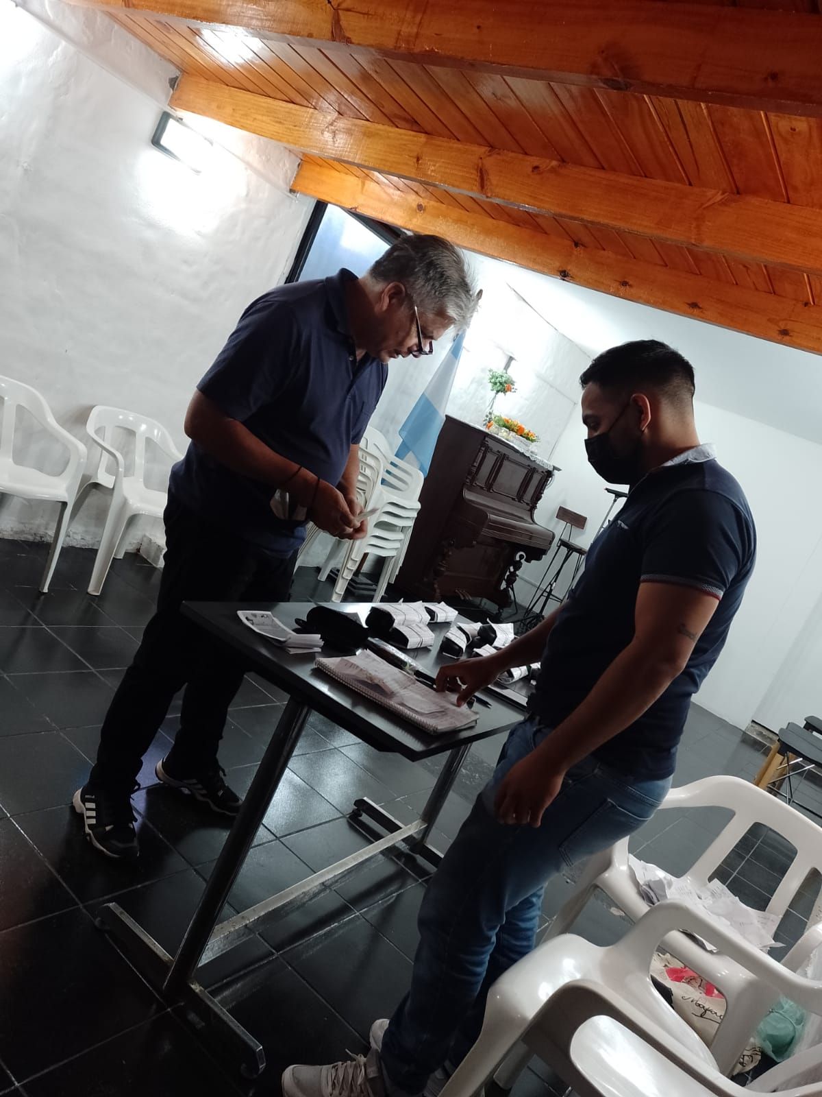 Dos hombres están de pie junto a una mesa con equipo en un pequeño taller interior.