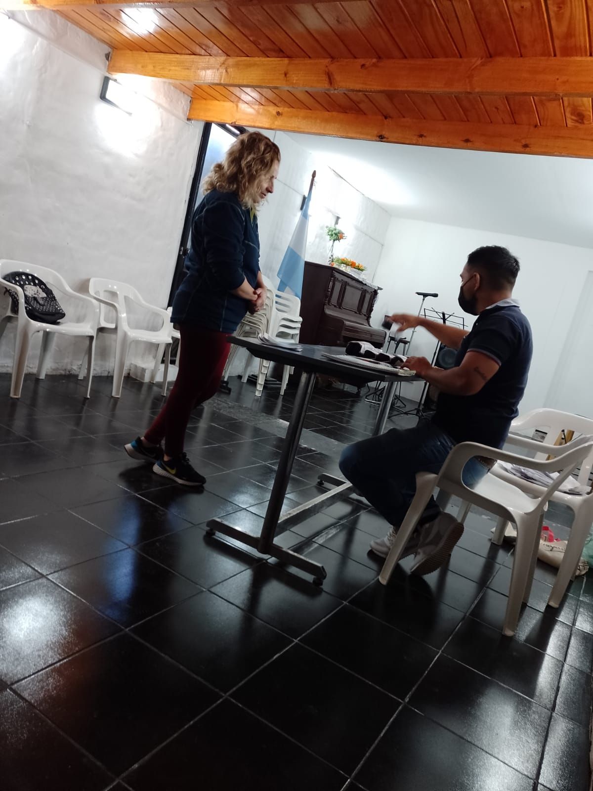 Dos personas están sentadas y de pie junto a una mesa en una habitación interior con suelo de baldosas, sillas de plástico blanco y techo de madera.