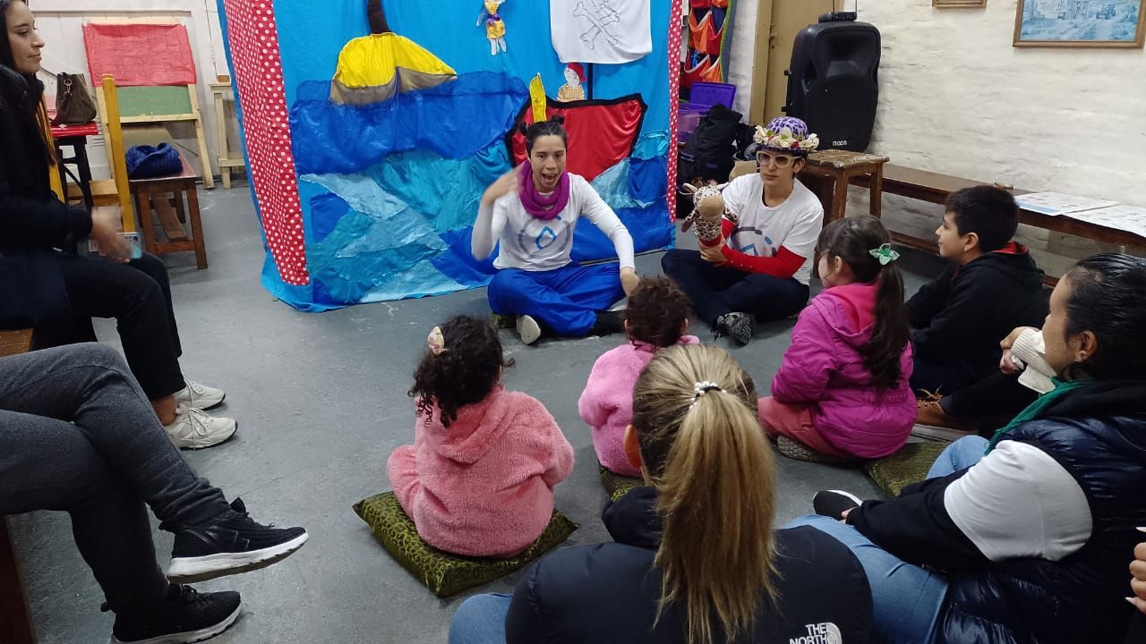 En un aula, los niños están sentados sobre esterillas mientras una mujer dirige una actividad frente a un fondo azul.