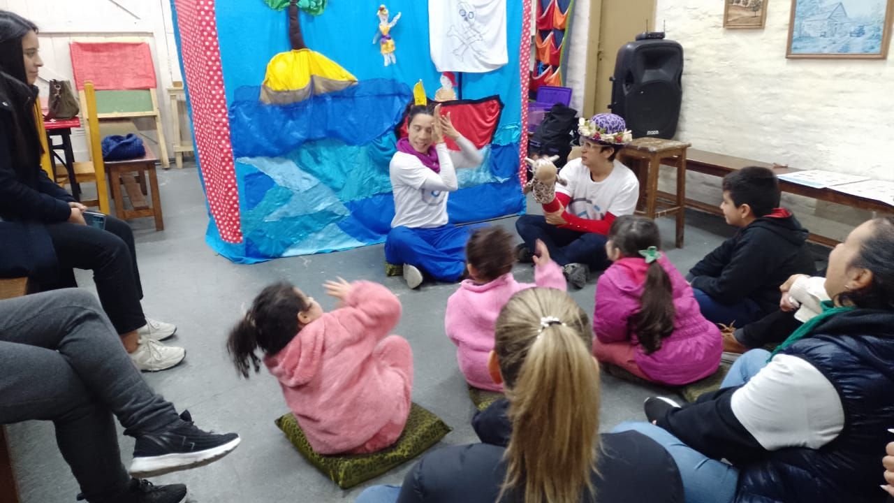 En un aula, unos niños sentados en el suelo observan a una persona que habla junto a una colorida pantalla azul.