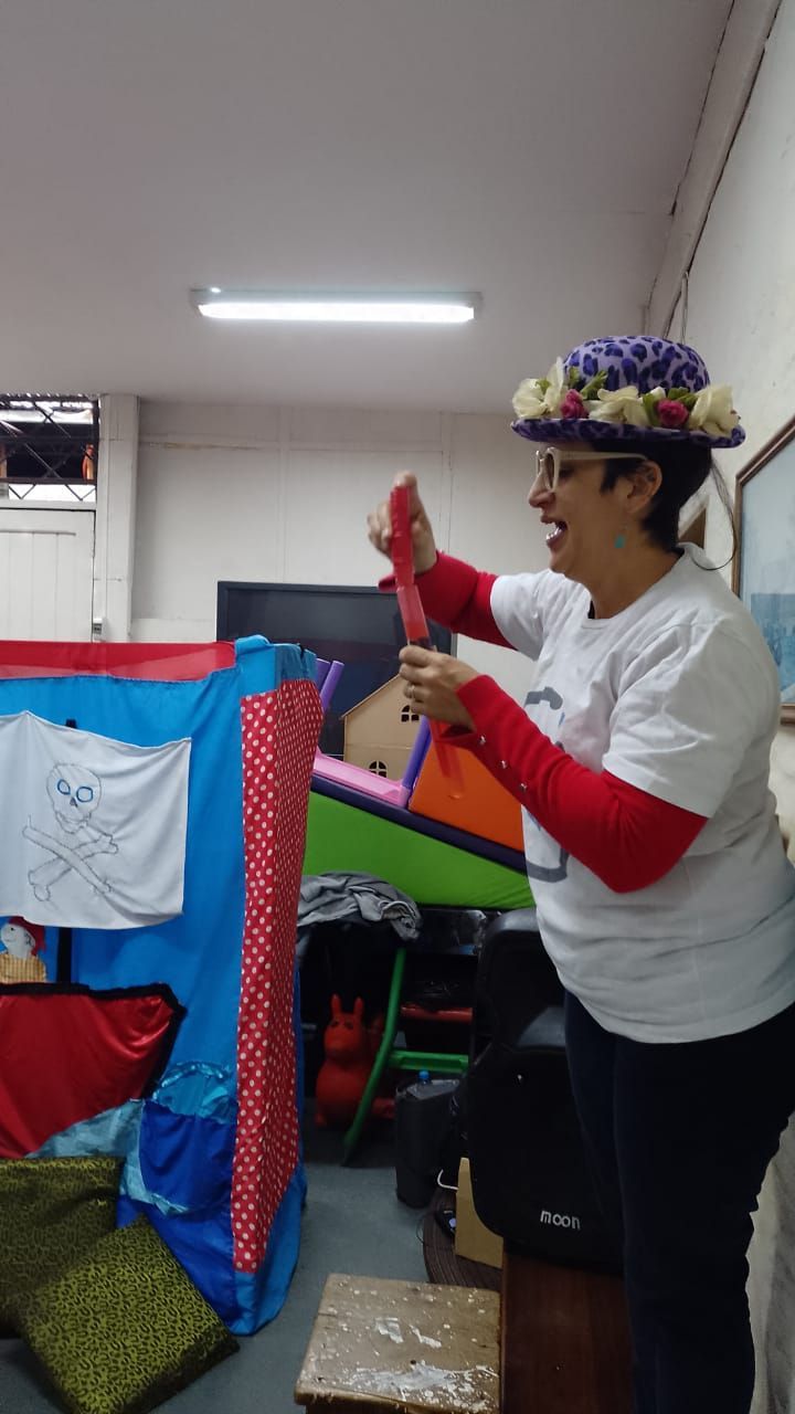 Una persona con un sombrero de flores sostiene un objeto rojo junto a una colorida decoración cubierta con tela en el interior de un edificio.