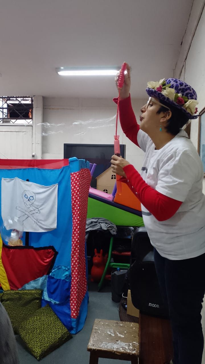 Mujer con camisa blanca y sombrero de flores estirando el brazo para ajustar una lámpara de techo junto a coloridas telas colgantes.