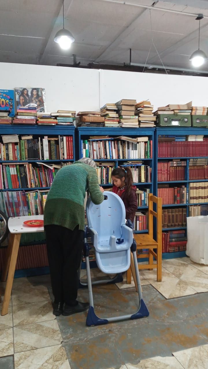 Una persona se inclina sobre un niño sentado en una biblioteca, con estanterías y libros apilados al fondo.
