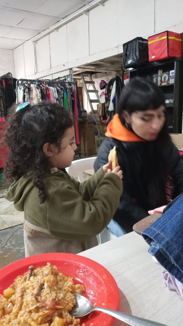 Un niño extiende la mano hacia una persona sentada en una mesa con un plato rojo de comida en una habitación interior.