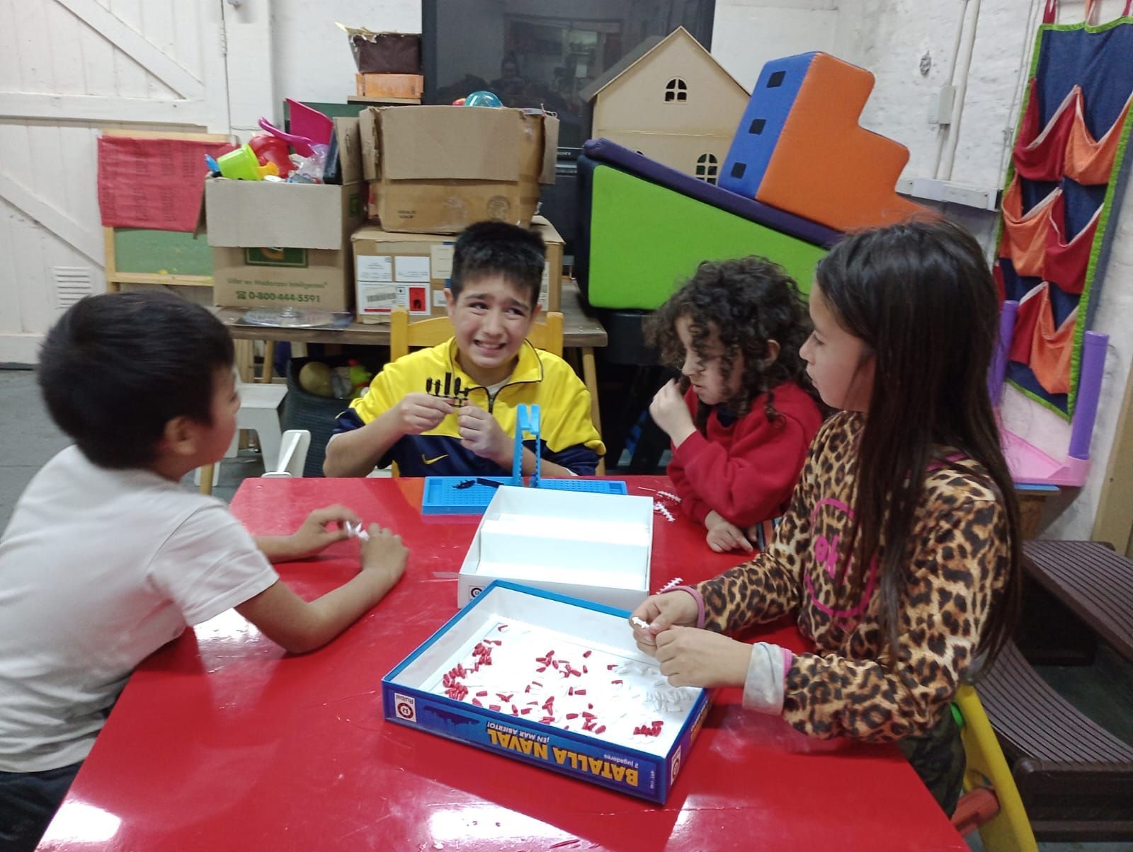 Niños jugando a un juego de mesa en una mesa roja en un aula colorida, sonriendo y concentrados en el juego.