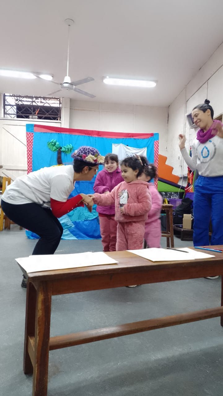 Dos niños y dos adultos en un aula; un adulto se inclina hacia un niño cerca de una mesa y un tablón de anuncios.