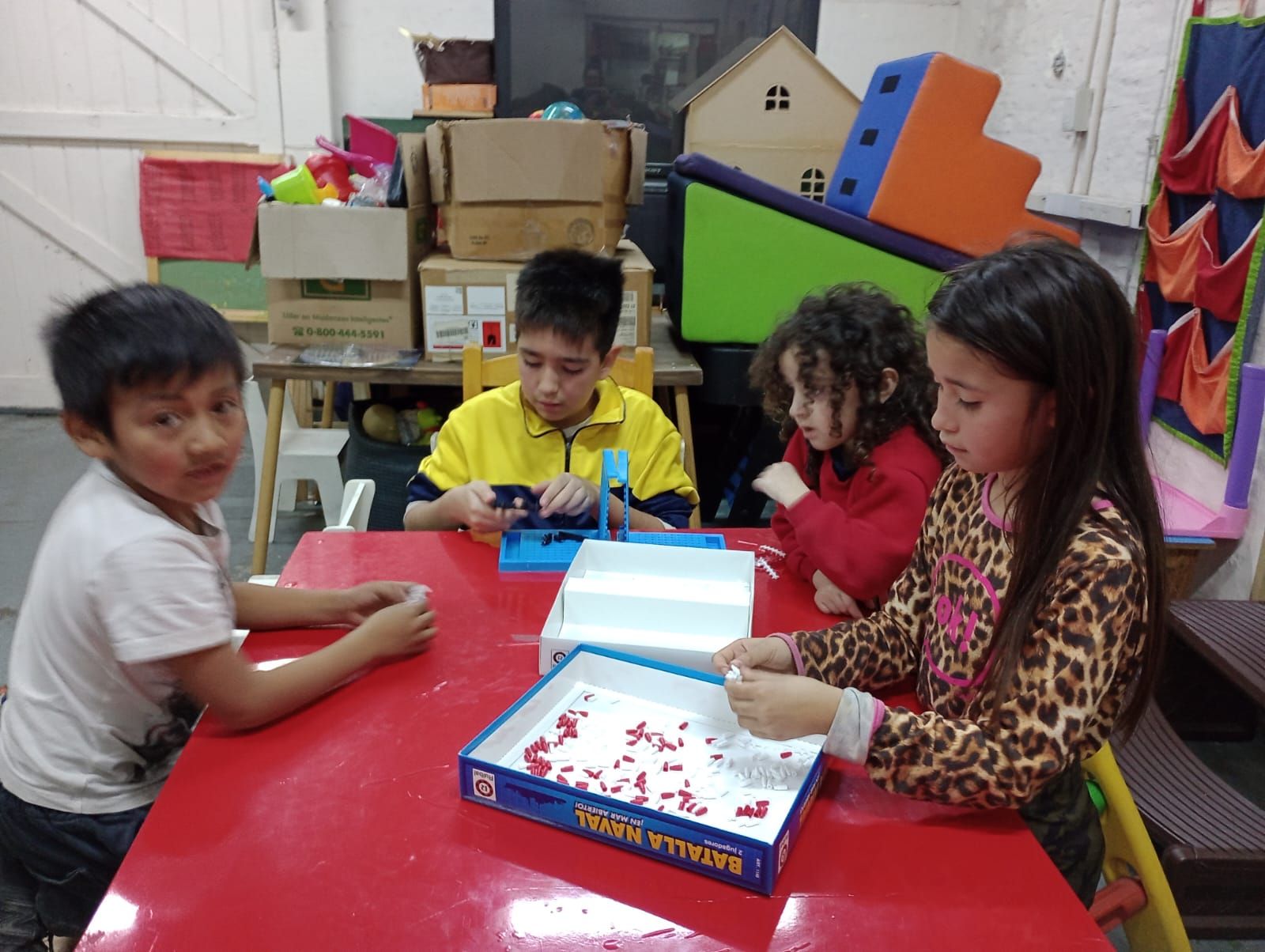 Cuatro niños están sentados alrededor de una mesa roja jugando a un juego de mesa en un aula colorida.