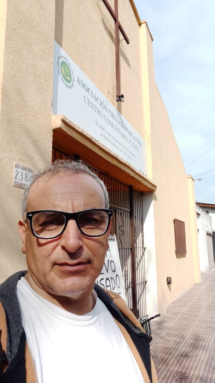Un hombre se toma una selfie en una calle soleada junto a un edificio beige con un letrero sobre la puerta.