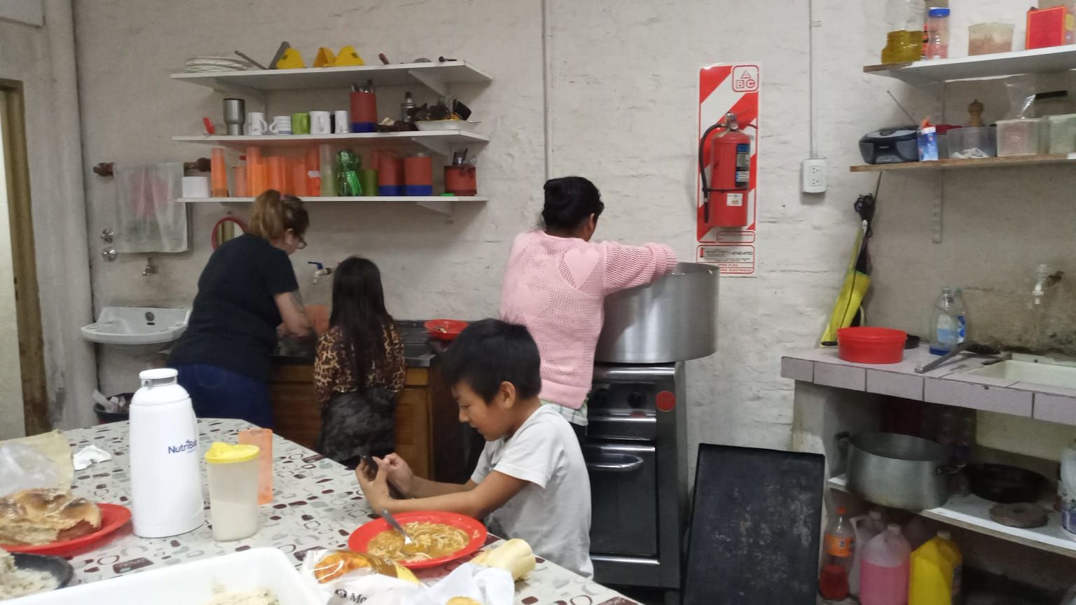 Personas preparando comida en una pequeña cocina con estantes, fregadero y un extintor rojo en la pared.