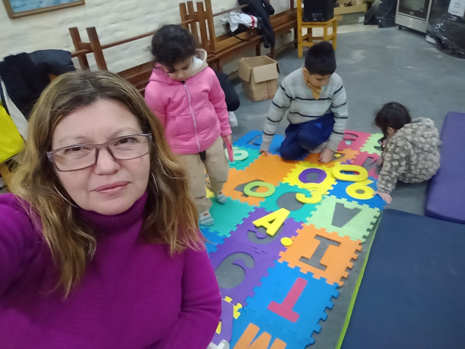 Mujer tomándose una selfie junto a niños sobre una colorida alfombra con el alfabeto en una habitación.