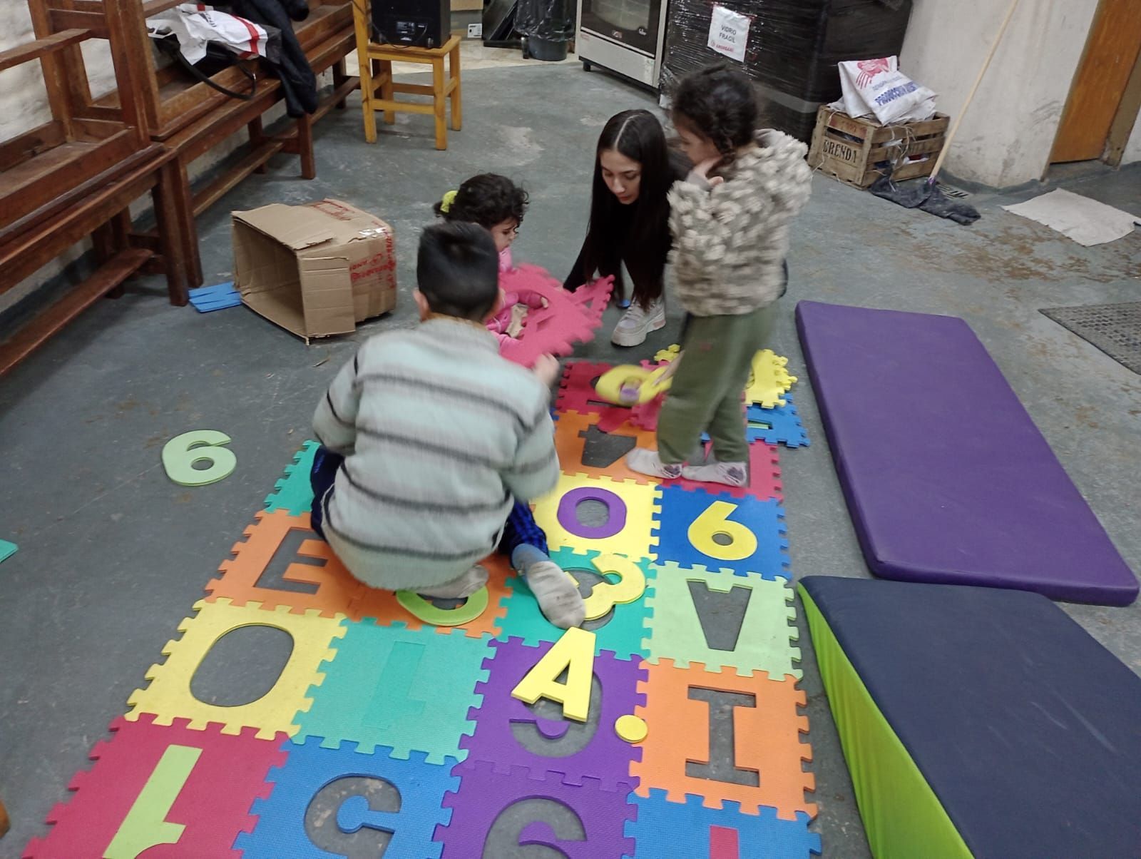 Niños sentados sobre una alfombra de números de colores, jugando con una mujer en una habitación.
