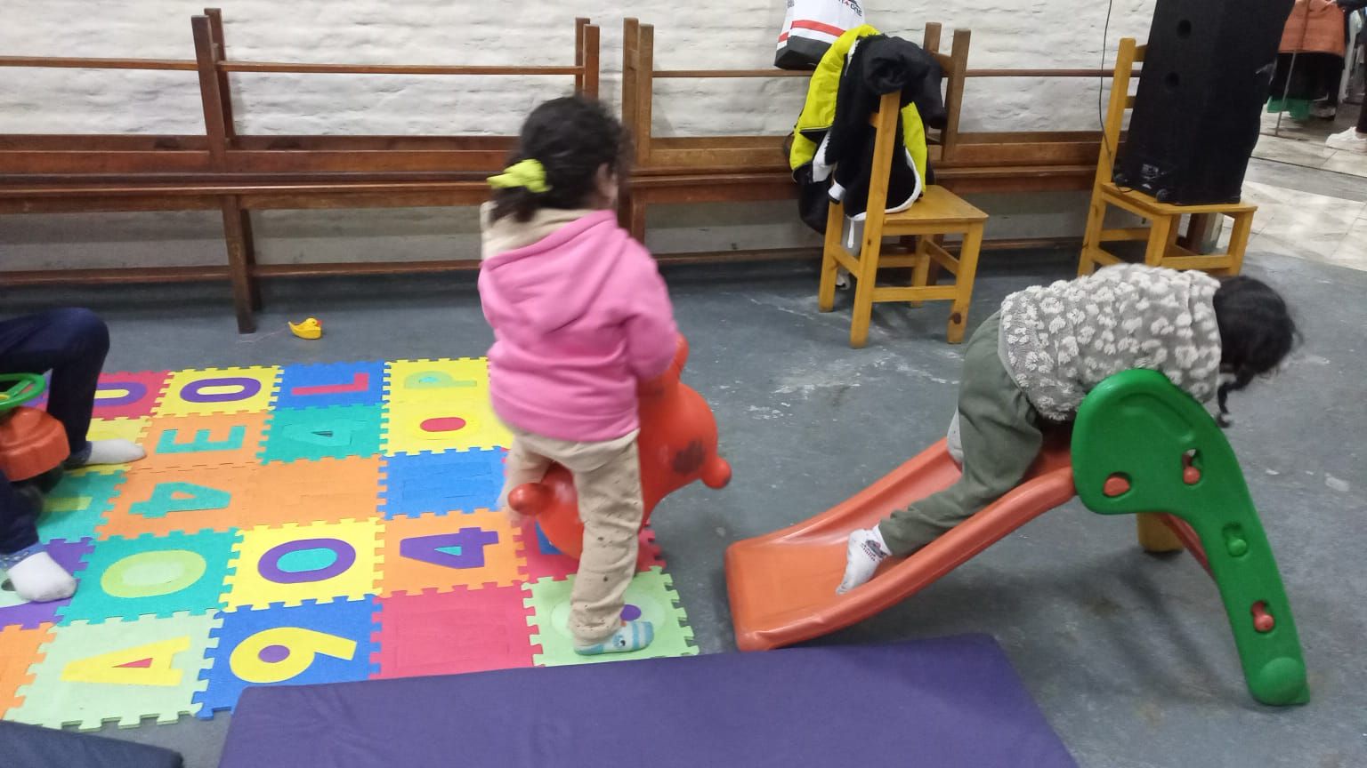 Niños jugando sobre coloridas colchonetas de espuma y un pequeño tobogán en un área de juegos interior.