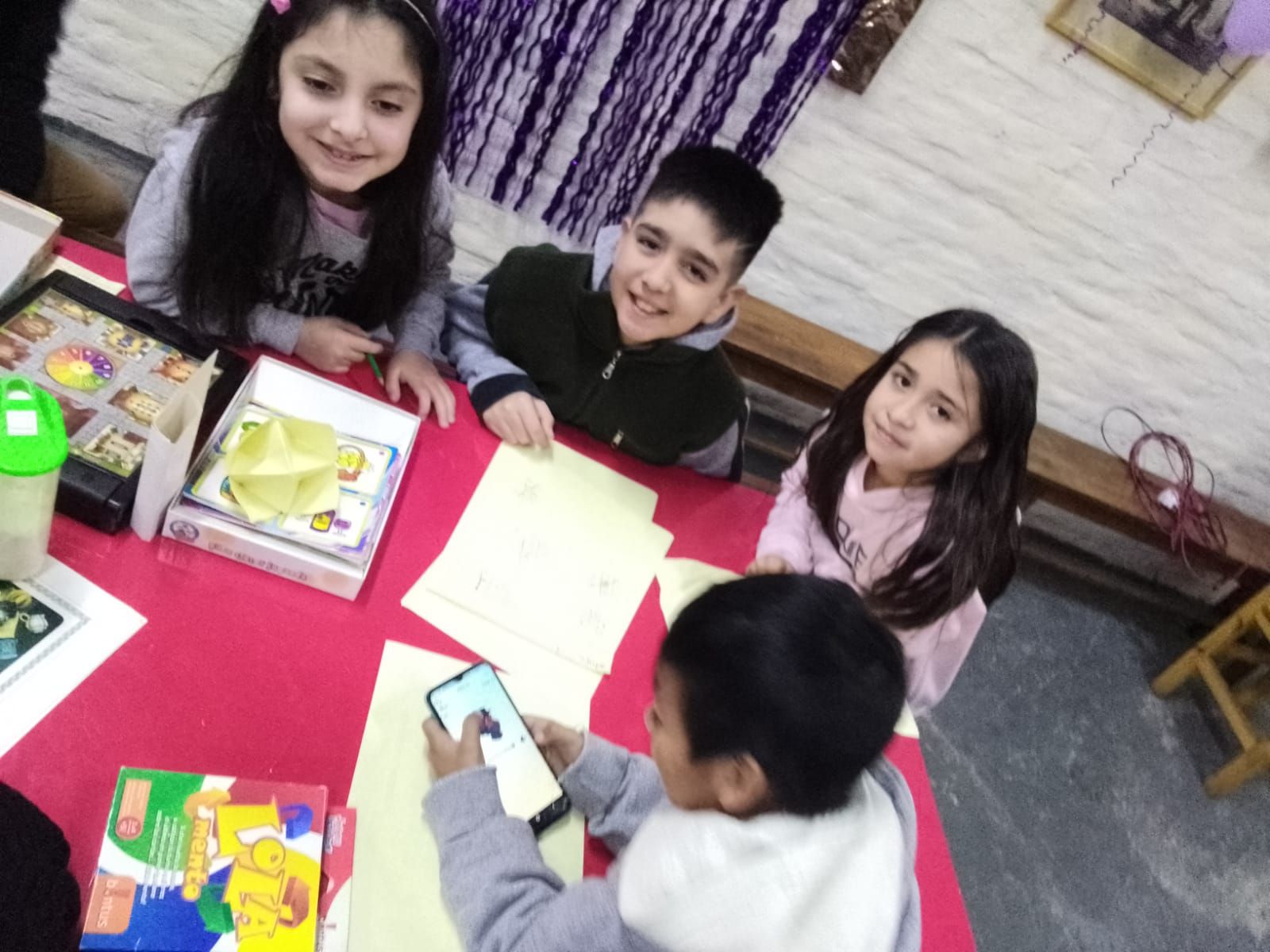 Cuatro niños están sentados alrededor de una mesa roja, jugando con juguetes y un teléfono inteligente dentro de la casa.