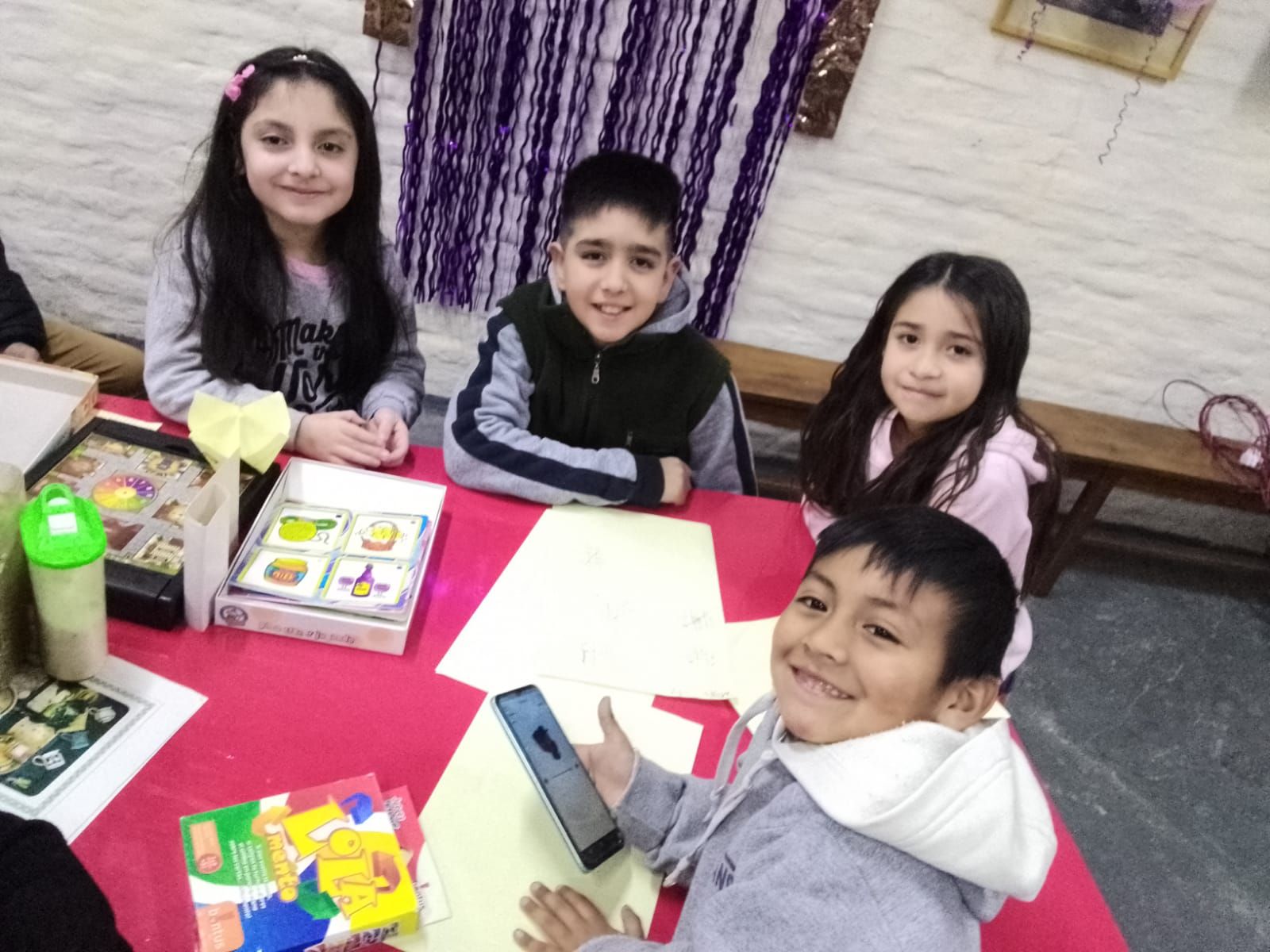 Cuatro niños están sentados alrededor de una mesa roja, sonriendo y sosteniendo un teléfono en una acogedora habitación con cortinas moradas.