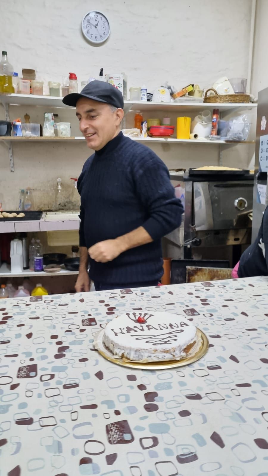 Una persona con suéter oscuro y gorra sonríe en una cocina junto a una mesa con un pastel decorado.