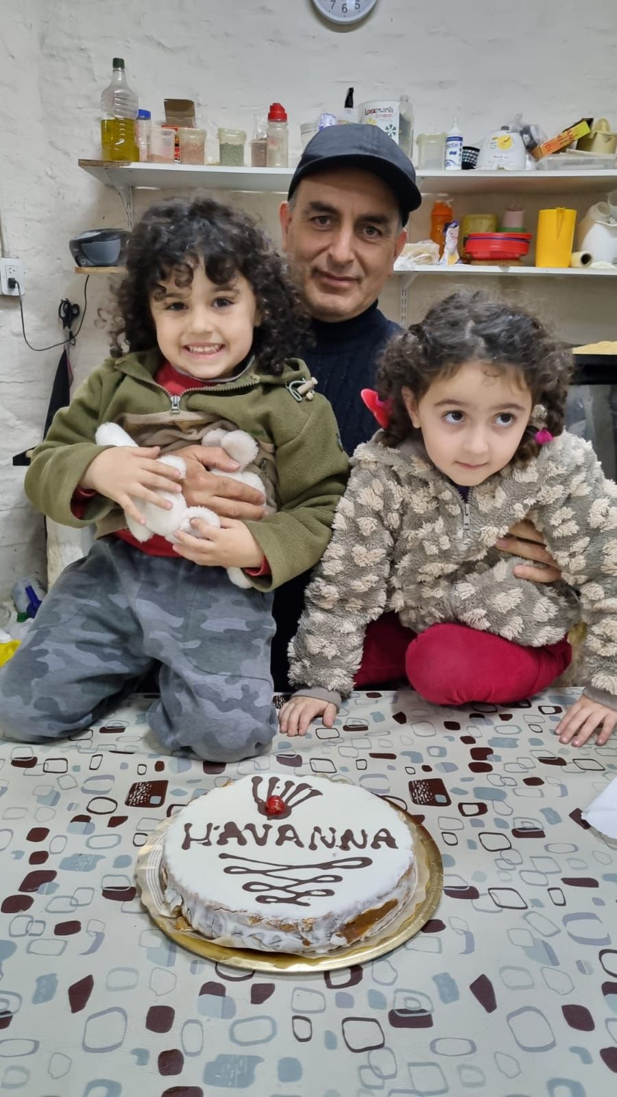 Un hombre sostiene a dos niños detrás de un pastel sobre una mesa en una acogedora cocina.