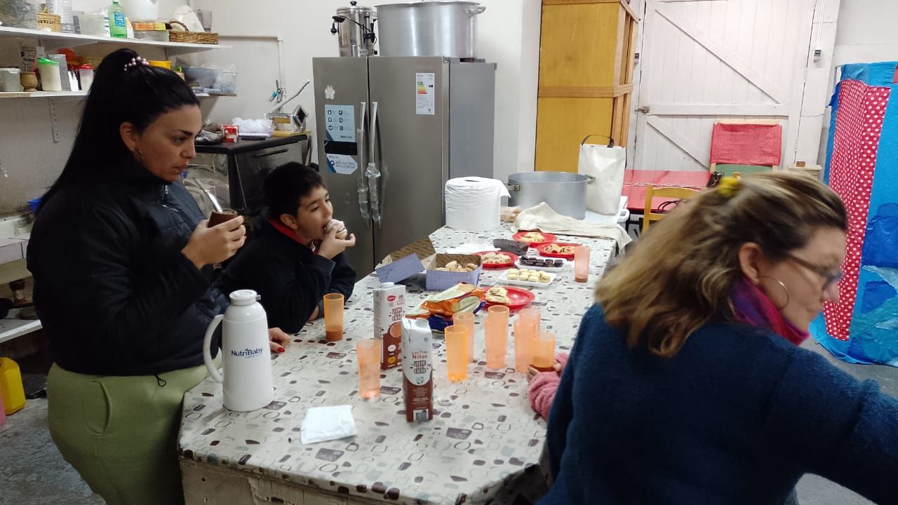 Tres personas están sentadas a una mesa en una pequeña cocina, comiendo y bebiendo con botellas y platos sobre un mantel de lunares.