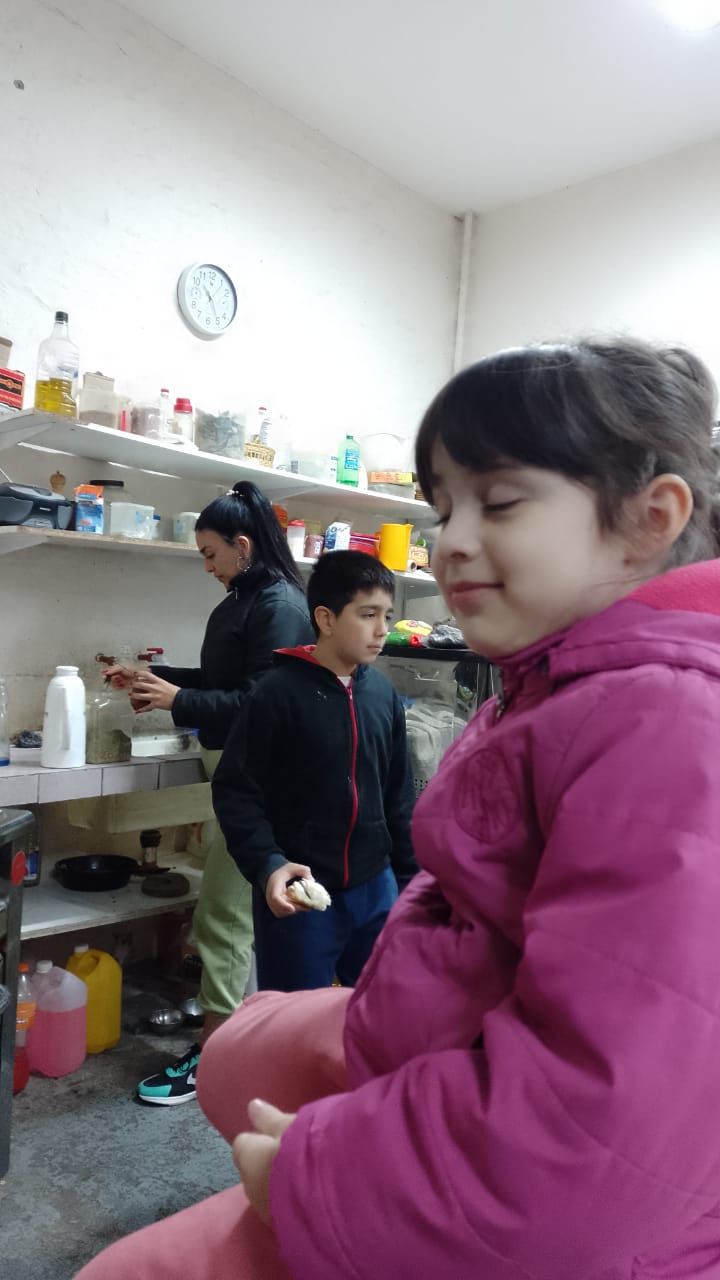 Dos niños en una cocina, uno con una chaqueta rosa en primer plano y el otro cocinando en la estufa.