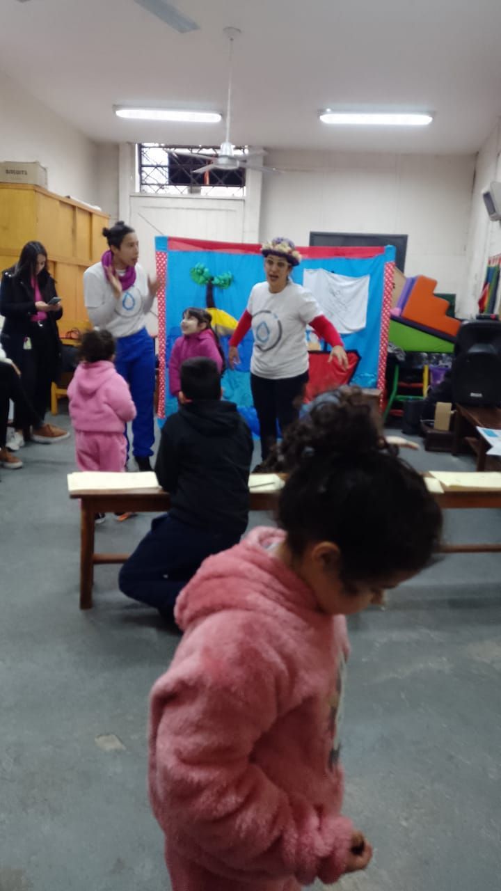 Actividad en el aula con niños de pie y sentados alrededor de un profesor cerca de un colorido panel informativo.