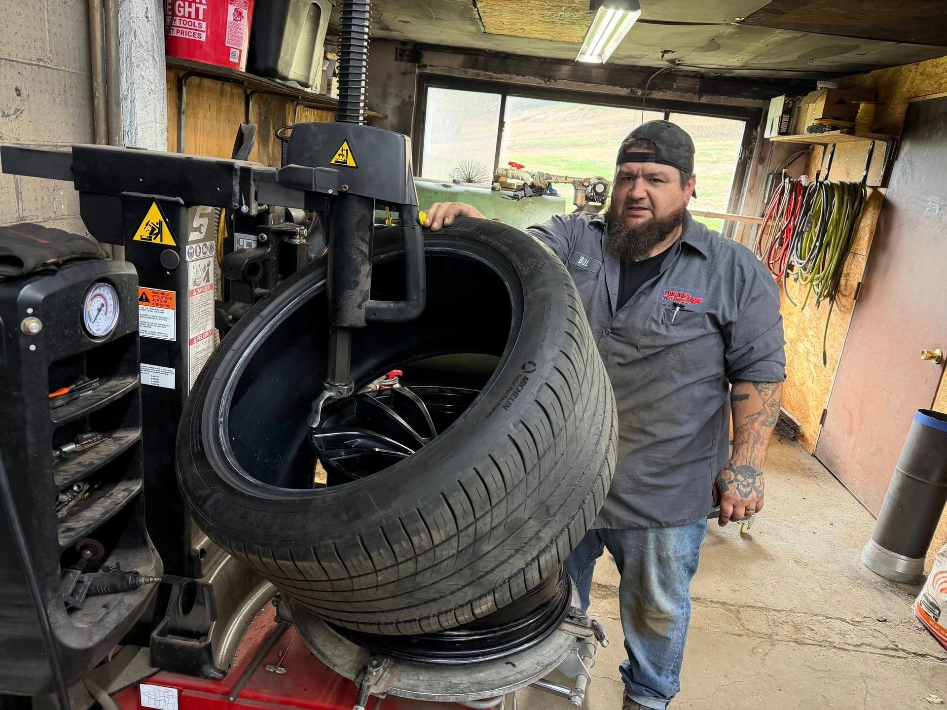 Man in a shop, holding a tire on a tire machine. He has a beard and is wearing a work shirt.