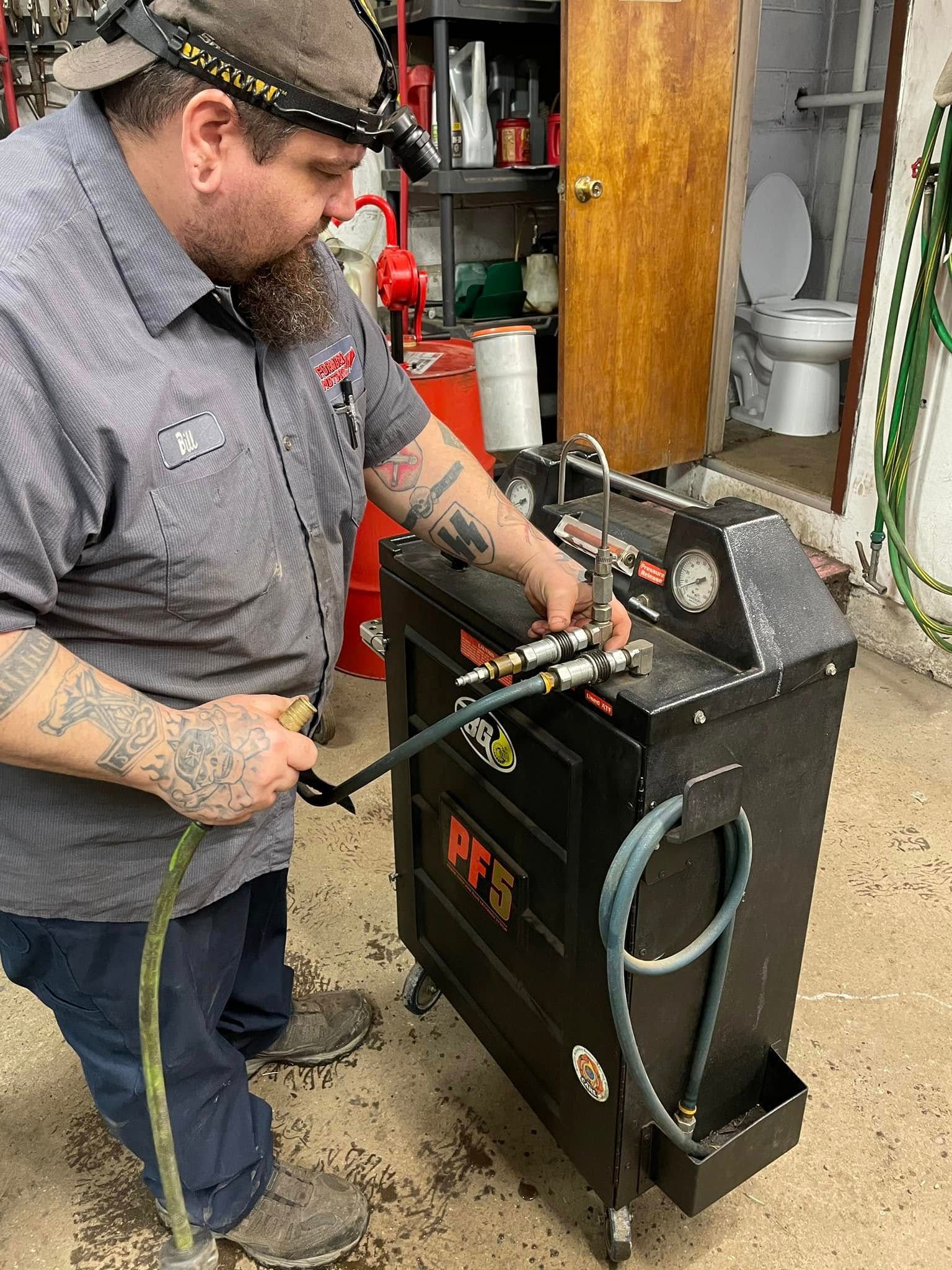 Mechanic working on a machine, possibly servicing a car. He wears a headlamp, holding a hose. Garage setting.