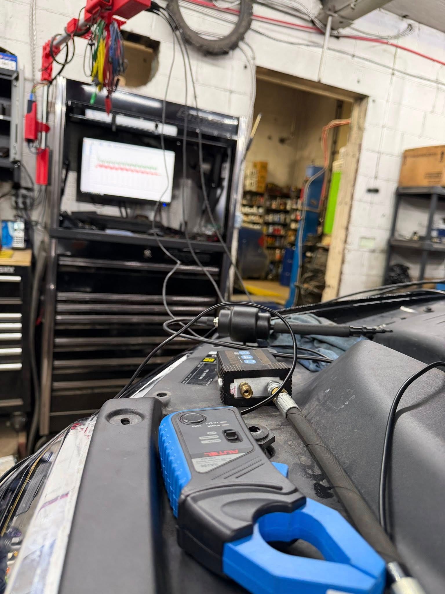 A black and blue automotive multimeter sits on a car hood, connected to equipment in a garage.
