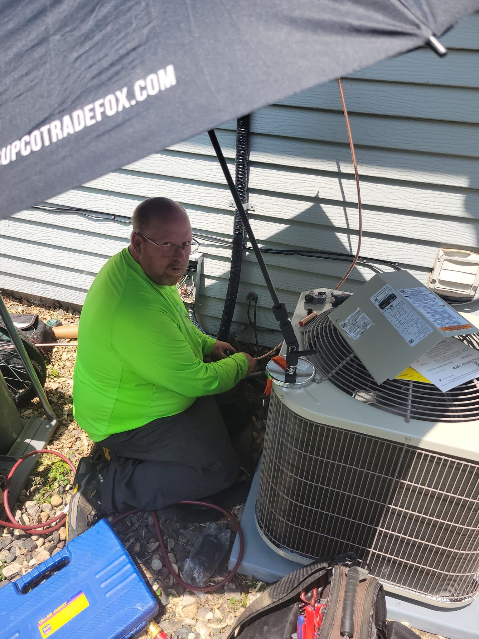A man is kneeling down next to an air conditioner under an umbrella.