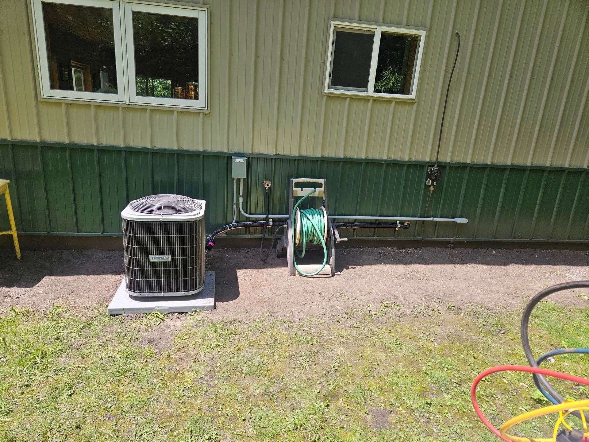 An air conditioner is sitting on the side of a building next to a hose reel.