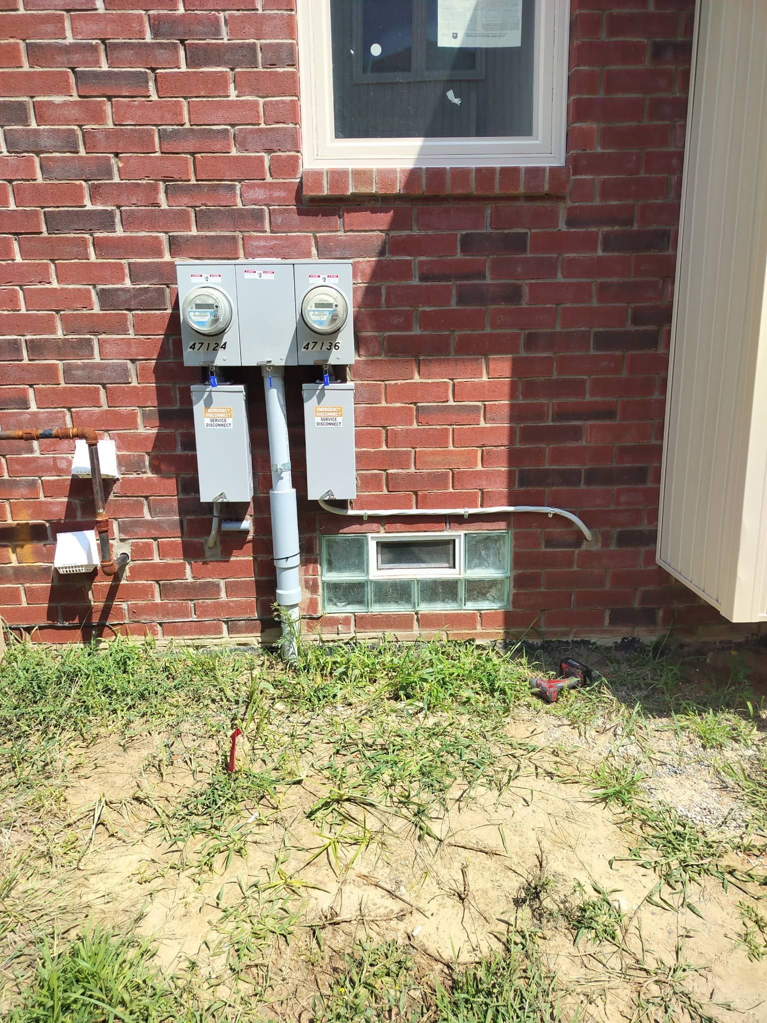 Two electric meters and utility boxes on a brick house wall above a small basement window and grassy yard