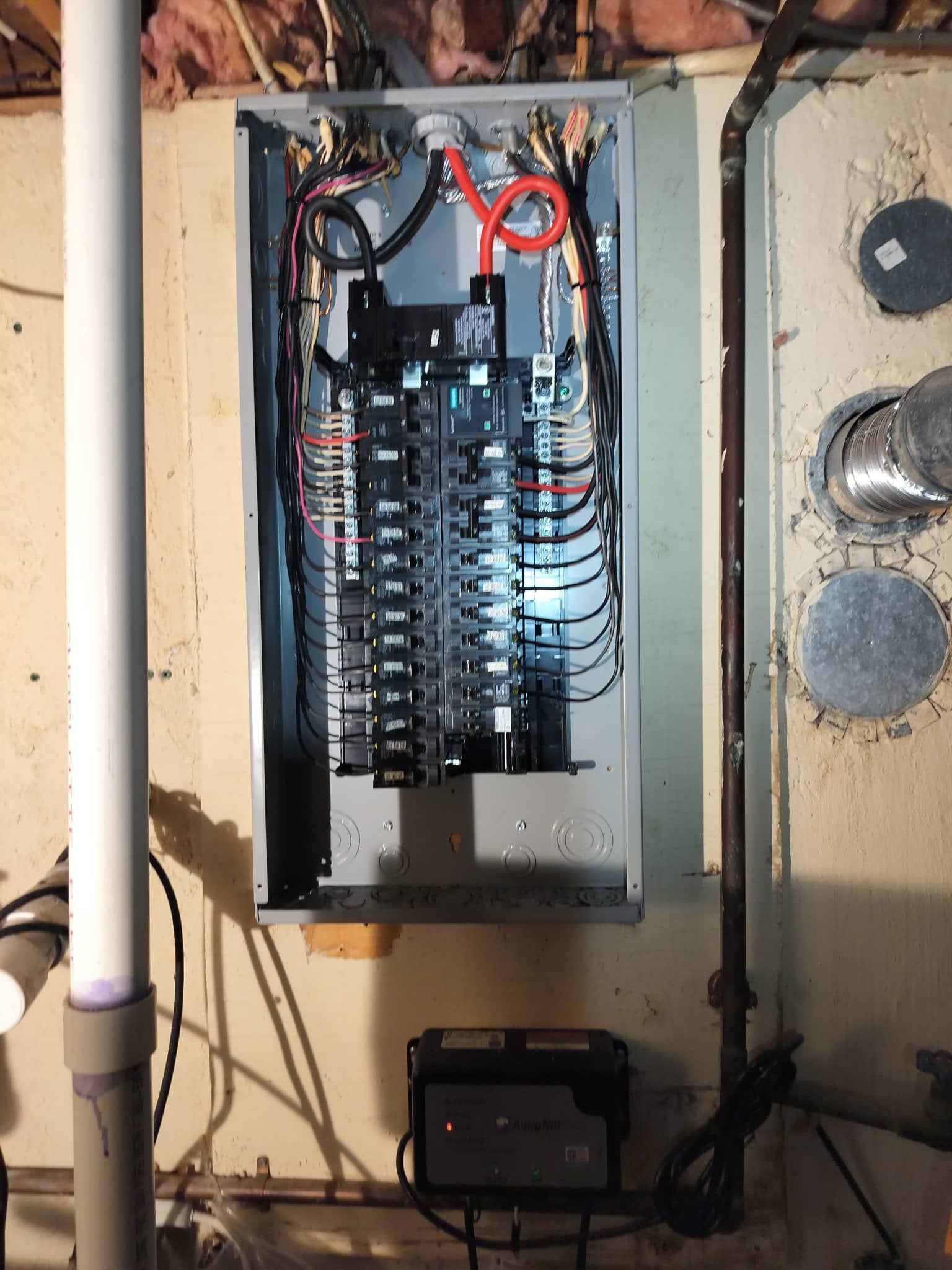 Electrical breaker panel with wiring in a wall recess, above a black device on the floor.