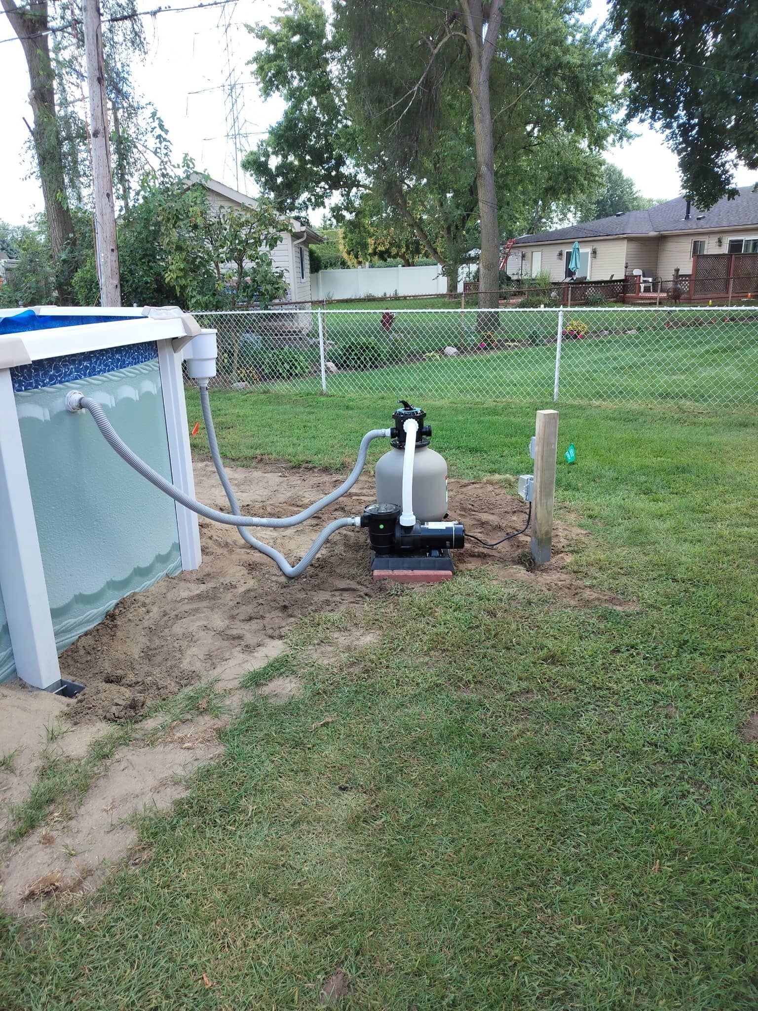 Outdoor pool pump setup beside an above-ground pool, with hoses and a fenced yard in the background