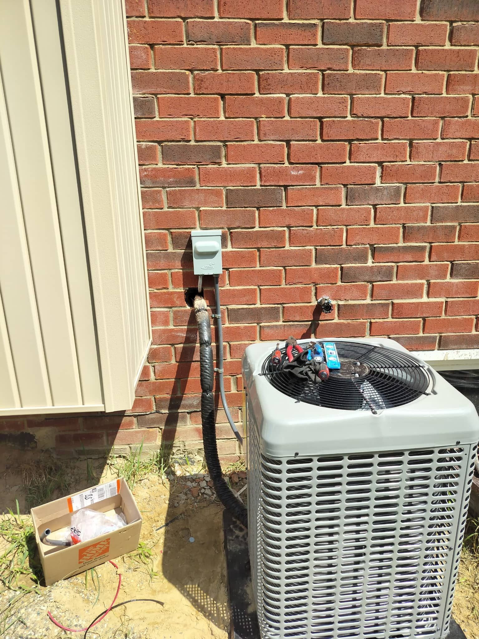 Outdoor air conditioner unit beside a brick wall with a disconnected black hose and open cardboard box nearby