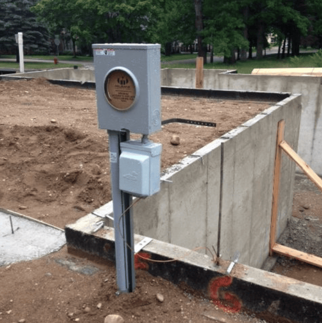 Gray utility pedestal beside a concrete foundation under construction, with dirt, wood braces, and a park background